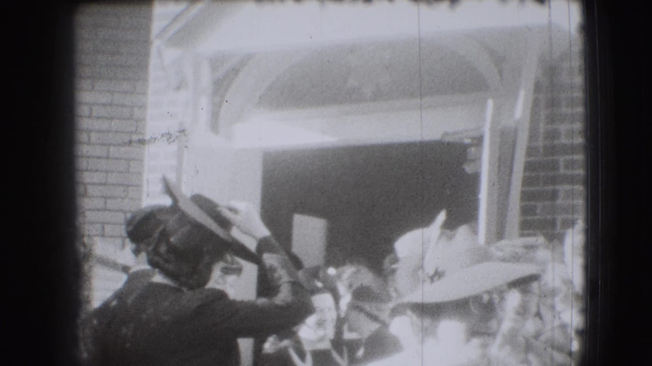 Women in hats gather near a brick building entrance as one figure lifts a hand toward her hat.