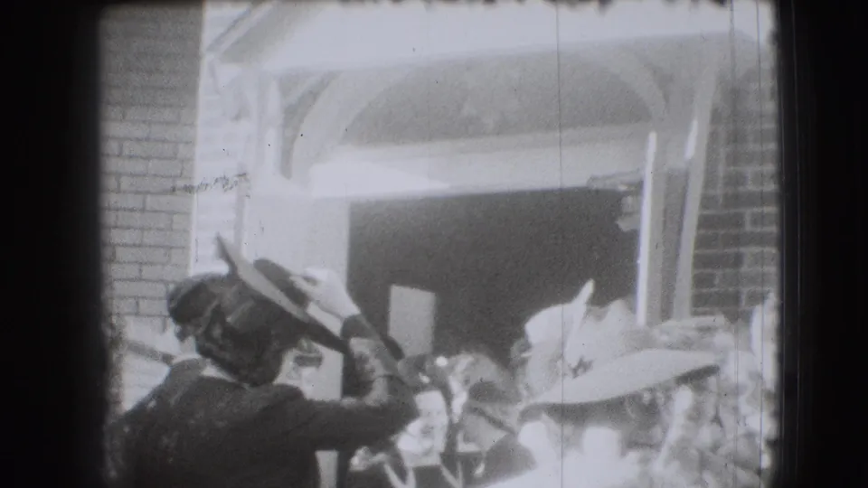 1941 Joliet home-movie frame showing women in vintage hats clustered at a brick arched doorway, with one woman adjusting her hat.