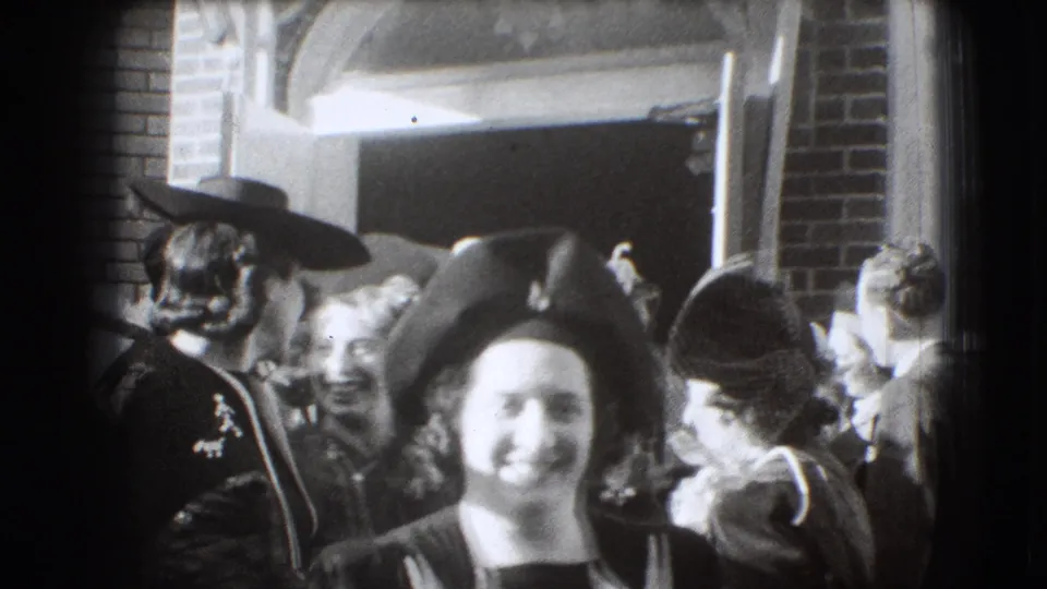 A woman in a dark brimmed hat smiles close to the camera, with other women gathered at the brick doorway in 1941 Joliet footage.