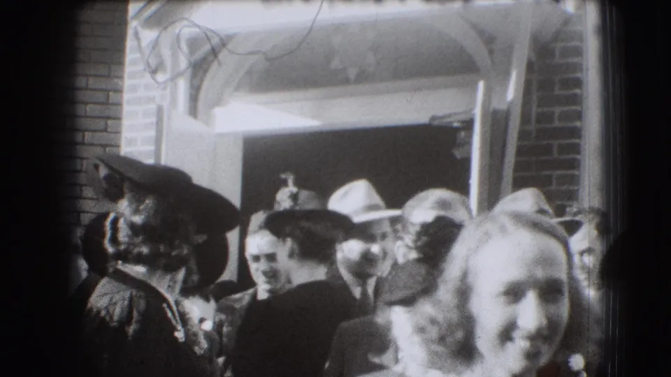 Adults in 1940s dress move past a brick building entrance in Joliet, with several faces visible close to the camera.