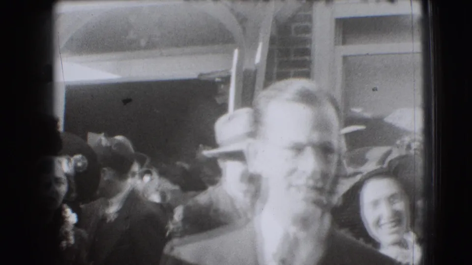 Soft 8mm frame from the Joliet crowd scene, with a blurred face in the foreground and hat-wearing adults behind.