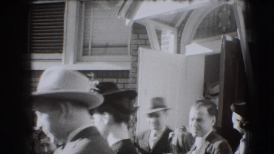 1941 Joliet frame with several adults in hats near a brick building entrance, including a man in the foreground and people by the doorway.