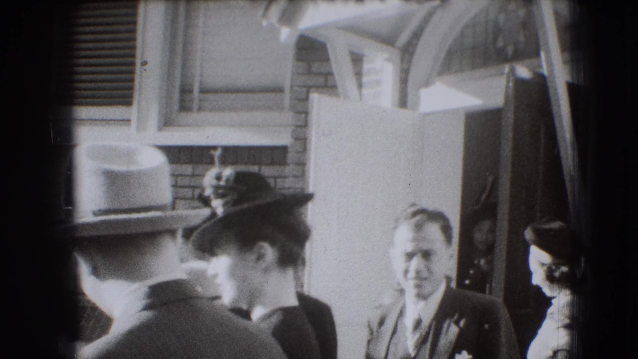 Adults in hats and suits stand outside a brick house near a porch.