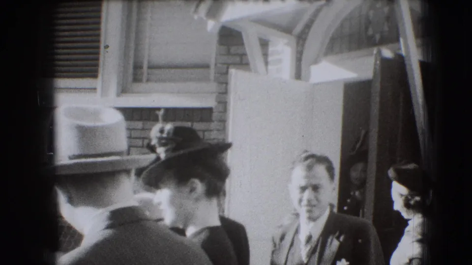 1941 Joliet home-movie frame showing adults in formal hats and suits gathered outside a brick residence.