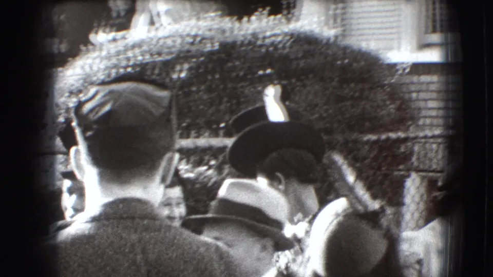 1941 Joliet outdoor gathering with adults in hats viewed from behind near a chain-link fence and shrubbery.