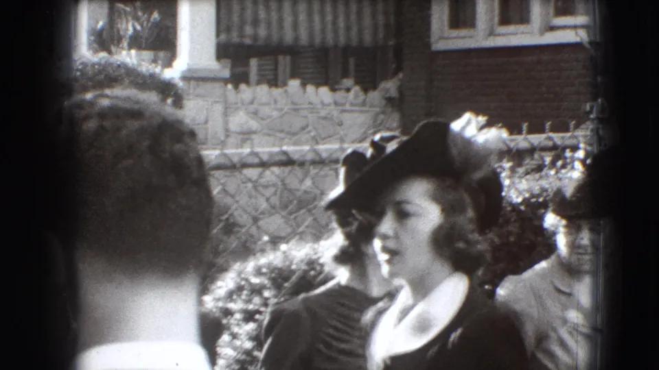 1941 Joliet frame showing a woman in a dark hat and light collar among adults outside a brick residential setting.