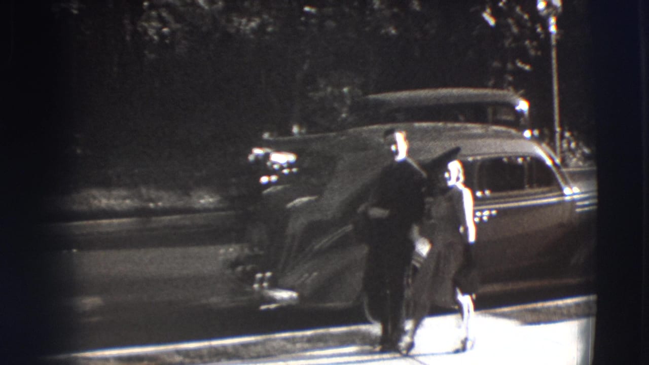 Man and woman walking on a sidewalk beside a rounded vintage car.