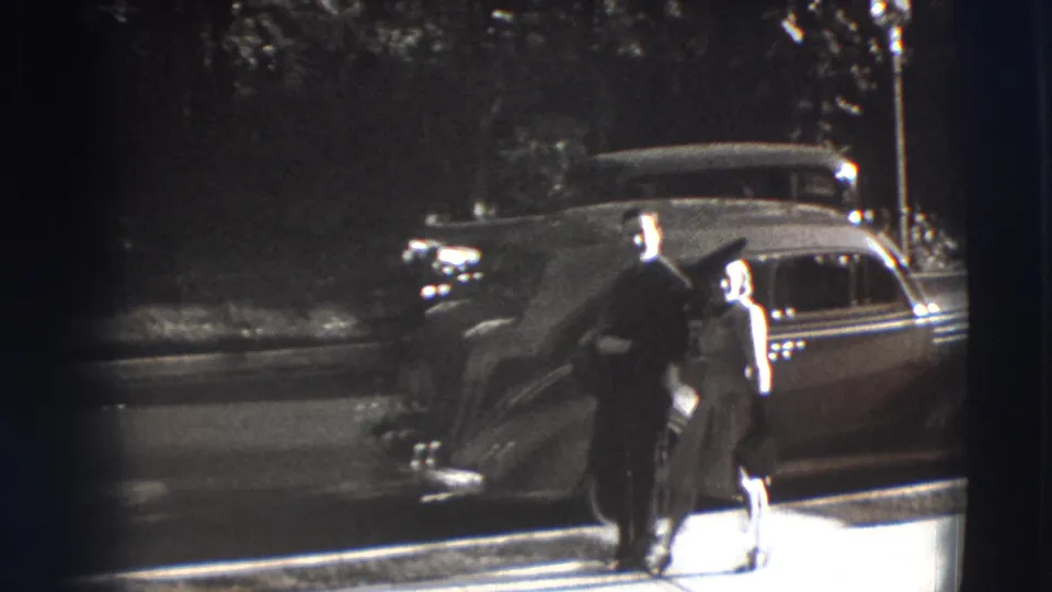 In 1941 Joliet, Illinois, a man and woman walk together on a sidewalk with a rounded vintage automobile behind them.