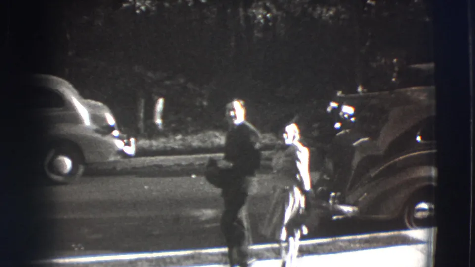 The couple continues along a Joliet sidewalk while rounded early 1940s automobiles appear along the street behind them.