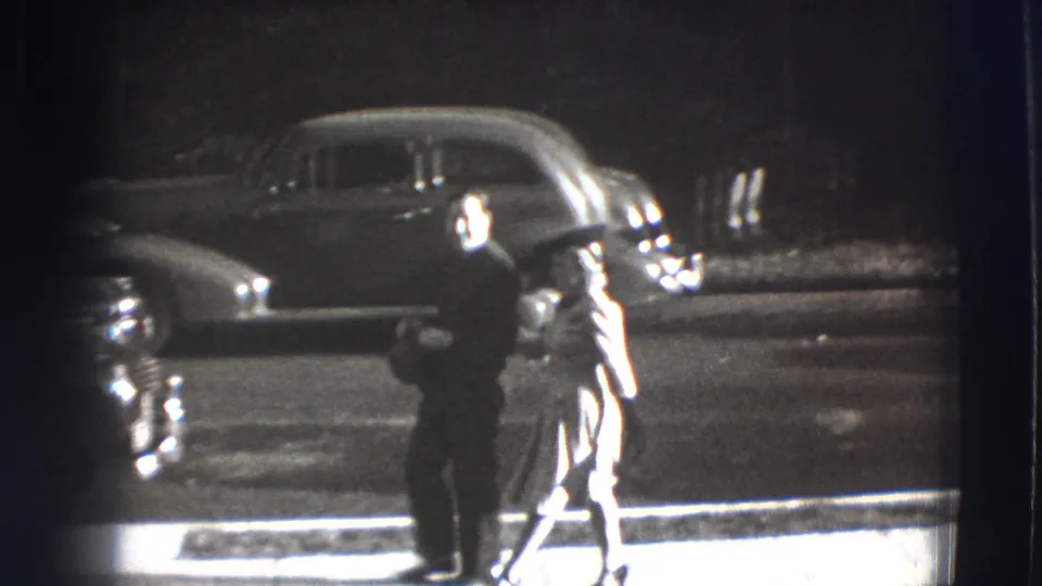 A man in dark clothing and a woman in a light dress walk beside the road in Joliet, with a rounded automobile in the background.