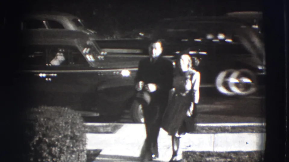 The couple approaches the camera on a Joliet sidewalk, framed by parked vintage cars and strong black-and-white contrast.