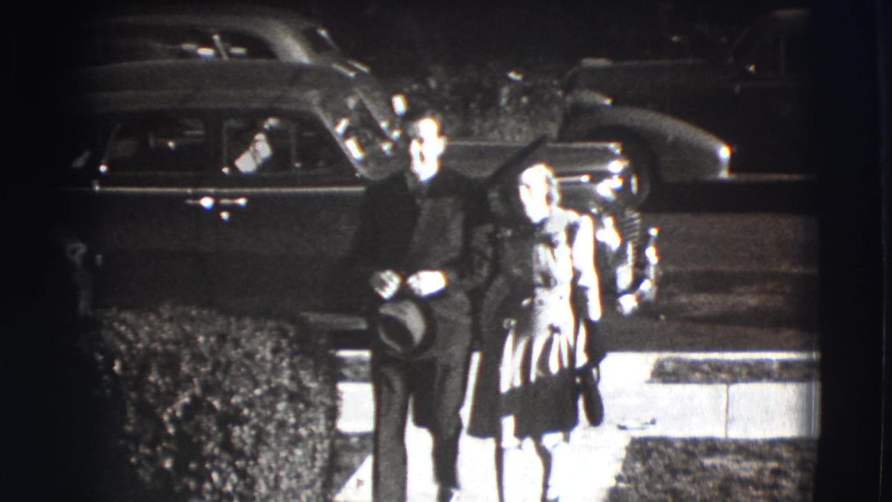 Man and woman walk from a street-side sidewalk toward the camera with parked cars behind them.