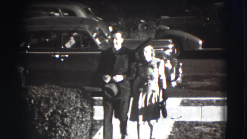 In a 1941 Joliet home-movie frame, a formally dressed man and woman approach the camera beside a hedge and parked 1940s cars.