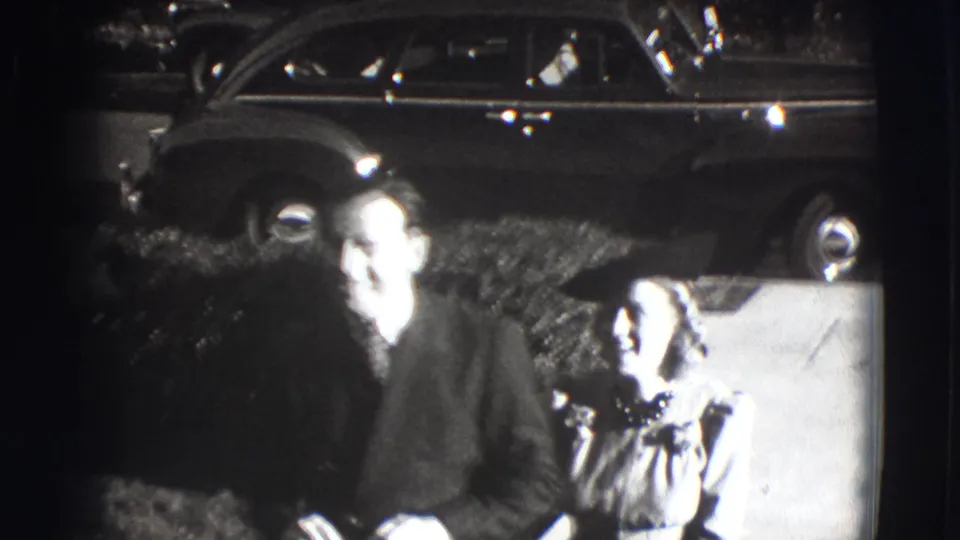 A close 8mm frame shows the man and woman near the lens, slightly soft from motion and exposure, with a parked car in the background.