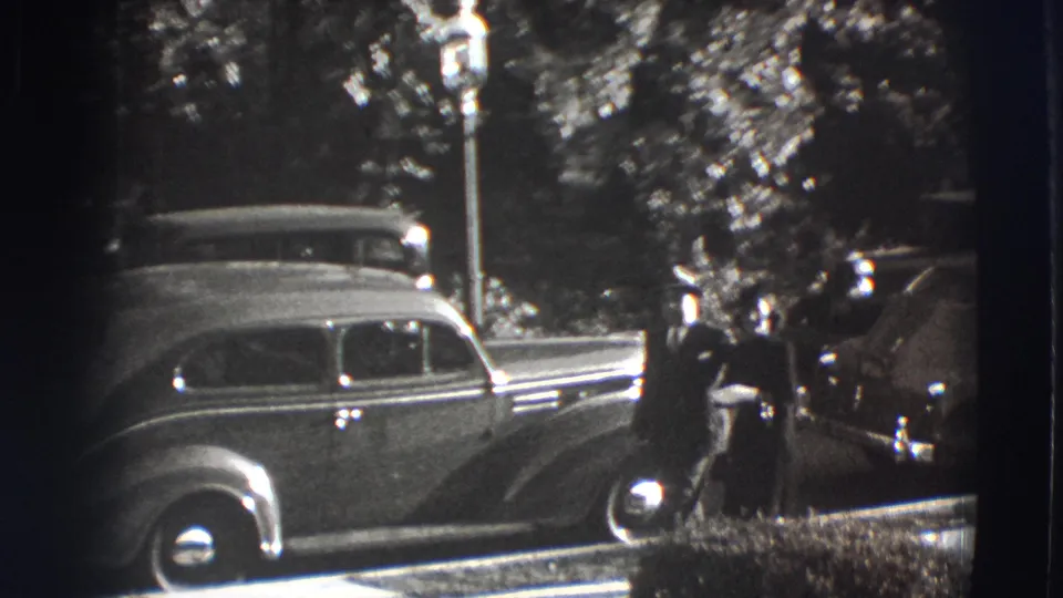 In 1941 Joliet, Illinois, a formally dressed couple appears beside a rounded sedan on a shaded sidewalk.