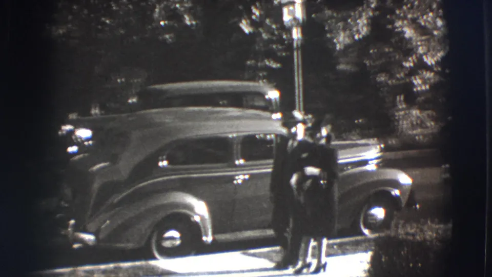 The couple walks arm in arm along a Joliet sidewalk, passing a 1940s-era sedan under trees.
