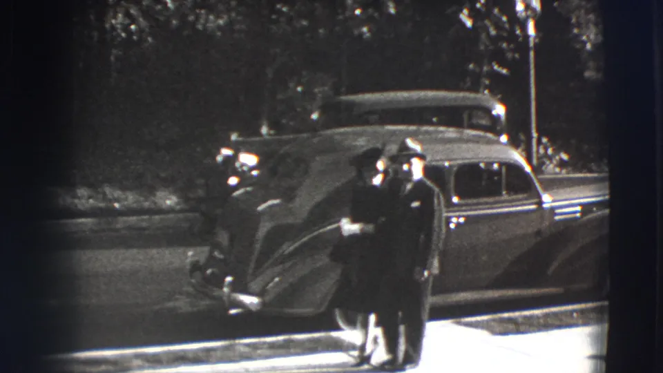 The man and woman approach the camera in formal 1940s clothing, with the sedan and shaded street behind them.