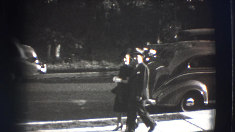 The couple moves along the sidewalk near the sedan, with trees and the street filling the background.
