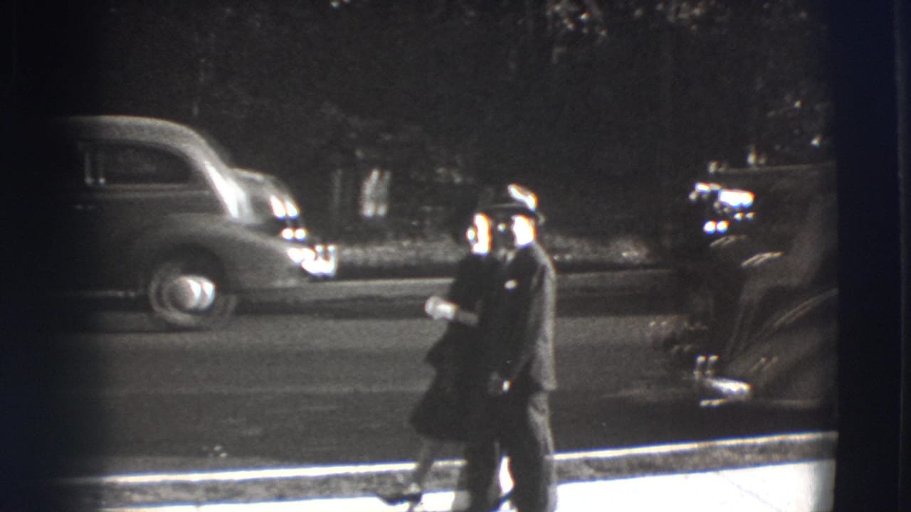 Man in a suit walks along a curb with parked 1940s cars behind him.