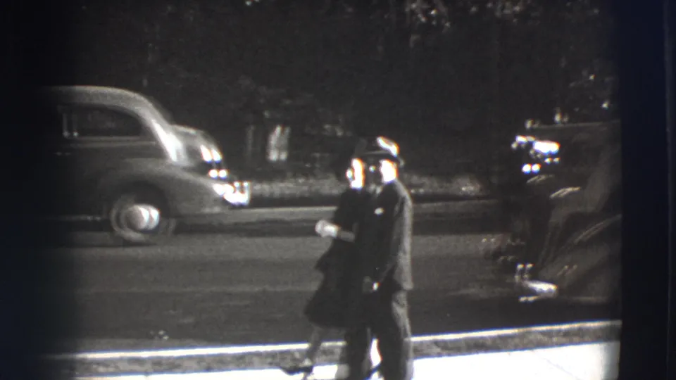 In the supplied 1941 Joliet home movie, a suited man walks near the curb as rounded automobiles sit parked along the street.