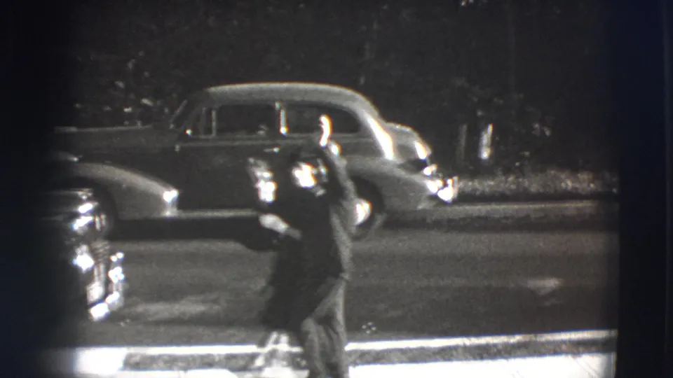 A woman in dark clothing moves across the curbside street scene, with mid-century automobiles visible behind her.