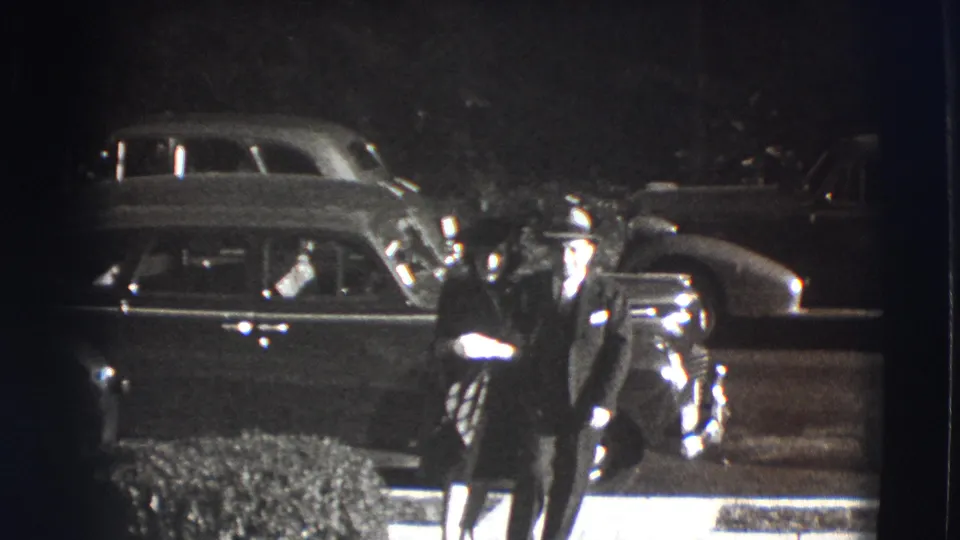 Two formally dressed adults appear near the curb beside a parked automobile in the 1941 Joliet home-movie scene.