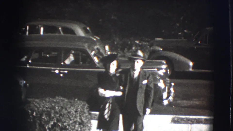 Two adults in formal 1940s clothing stand close together near shrubs and parked automobiles in the Joliet street scene.