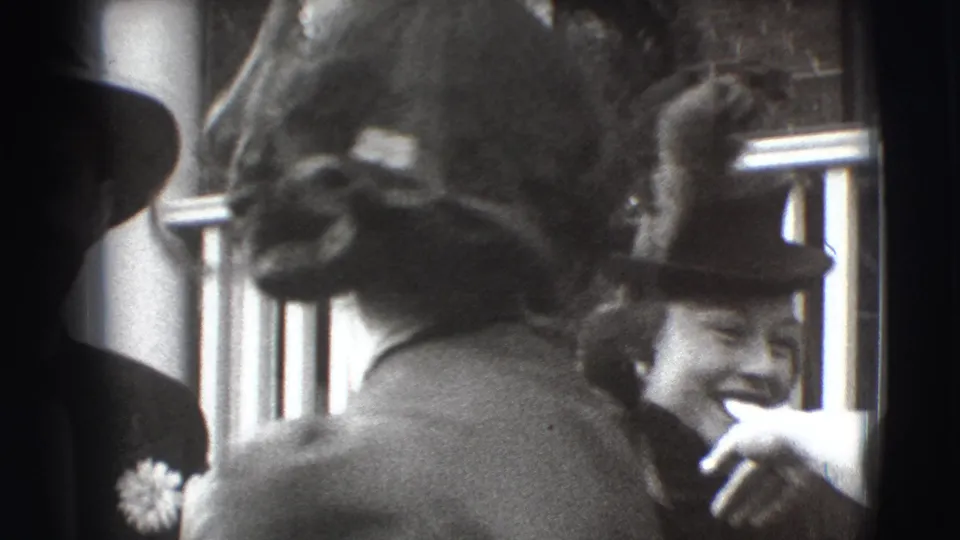 Black-and-white 1941 Joliet home-movie frame with guests in formal hats gathered outside a home near a porch railing.