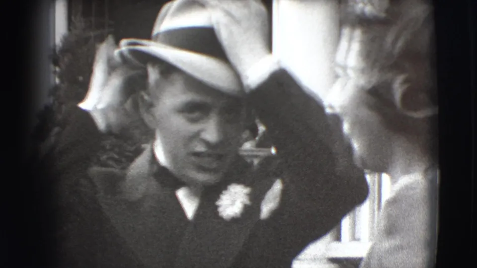 A suited man in a fedora adjusts his hat beside a woman during a 1941 Joliet exterior social gathering.