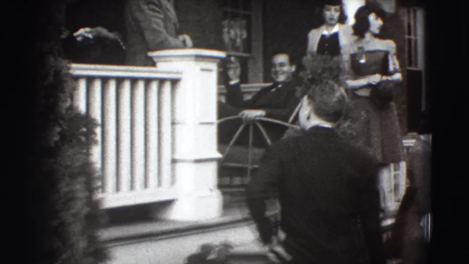 Several formally dressed guests gather around a Joliet porch, with women standing near the railing and men below.