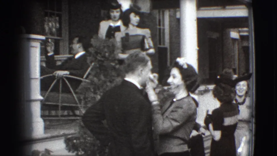 1941 Joliet home-movie frame of guests in suits, dresses, and hats talking outside a home porch.