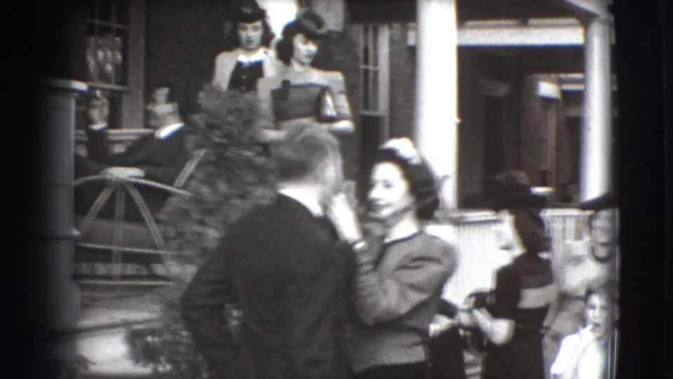 In 1941 Joliet home-movie footage, a woman grooms a man's face on a residential porch as onlookers in period clothing gather nearby.