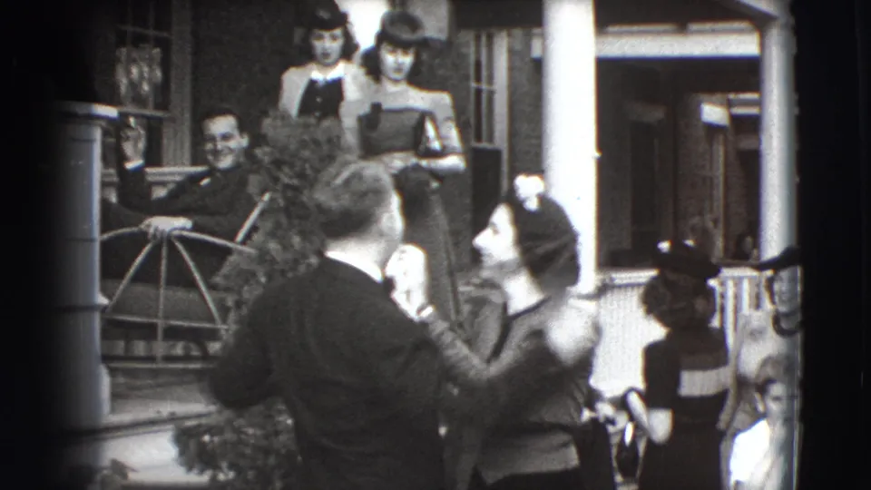 The man faces partly toward camera while a woman continues grooming his face, with adults in hats and dresses visible on the porch.