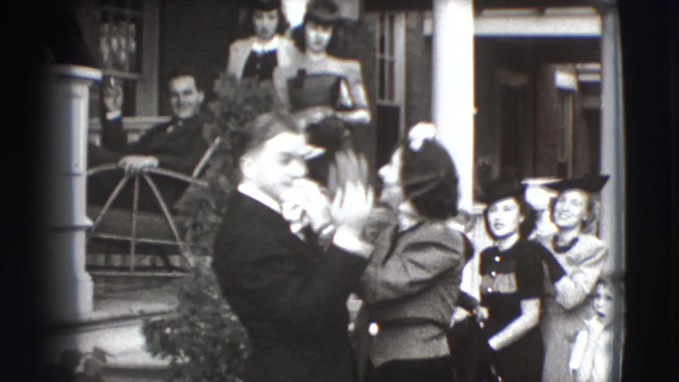 A woman in a hat lifts her hand near the suited man's face as porch onlookers, including women in hats and dresses, look toward the pair.