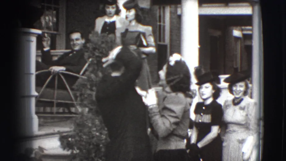 The suited man raises his hand to fix his hair as the woman remains beside him and several porch onlookers in 1940s-style clothing watch.