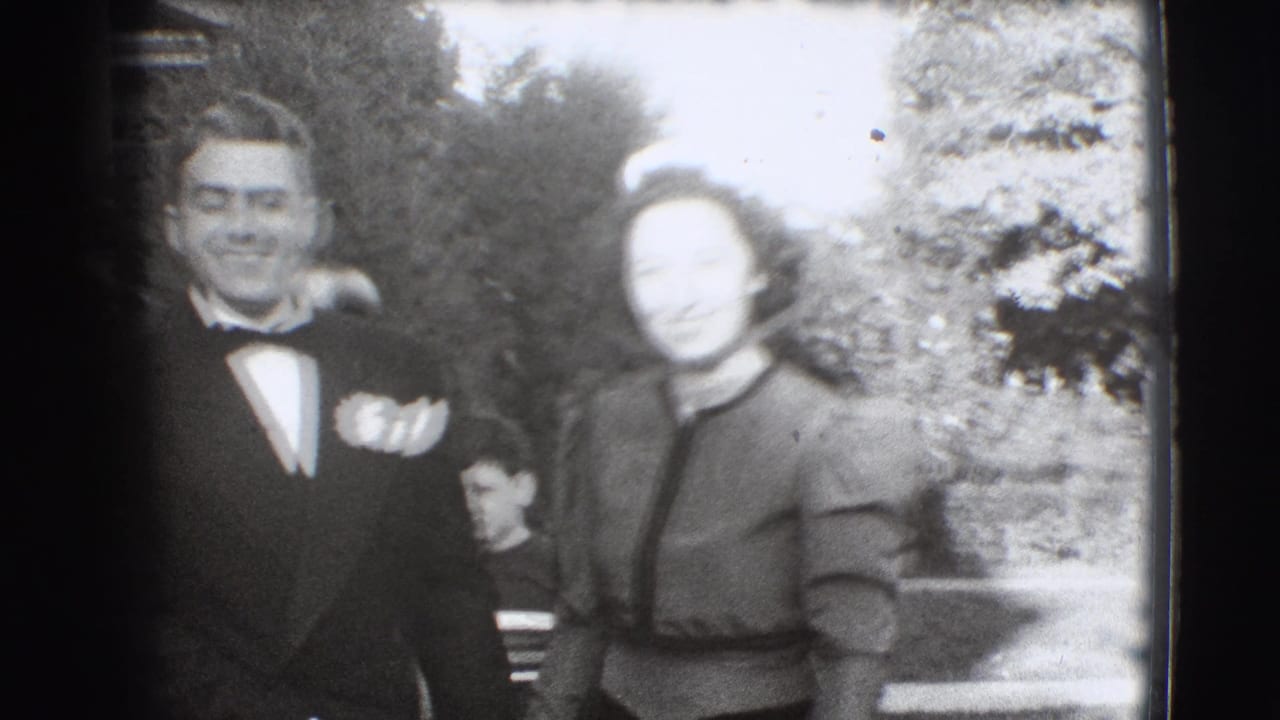 Young man in tuxedo and woman in formal dress pose outdoors beside shrubbery.