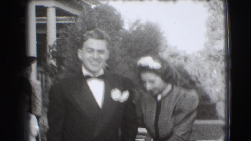 A 1941 Joliet home-movie frame shows a smiling man in tuxedo beside a woman in formal dress and headpiece near a porch.