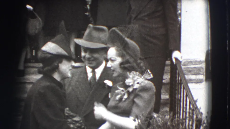 In 1941 Joliet, Illinois home-movie footage, adults in suits, dresses, and hats stand together beside an outdoor staircase.