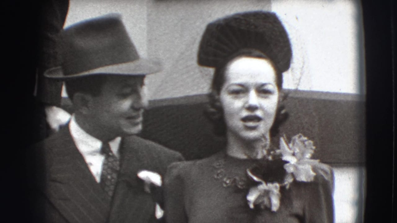 Man in a fedora and suit smiles beside a woman in a decorative hat and corsage.