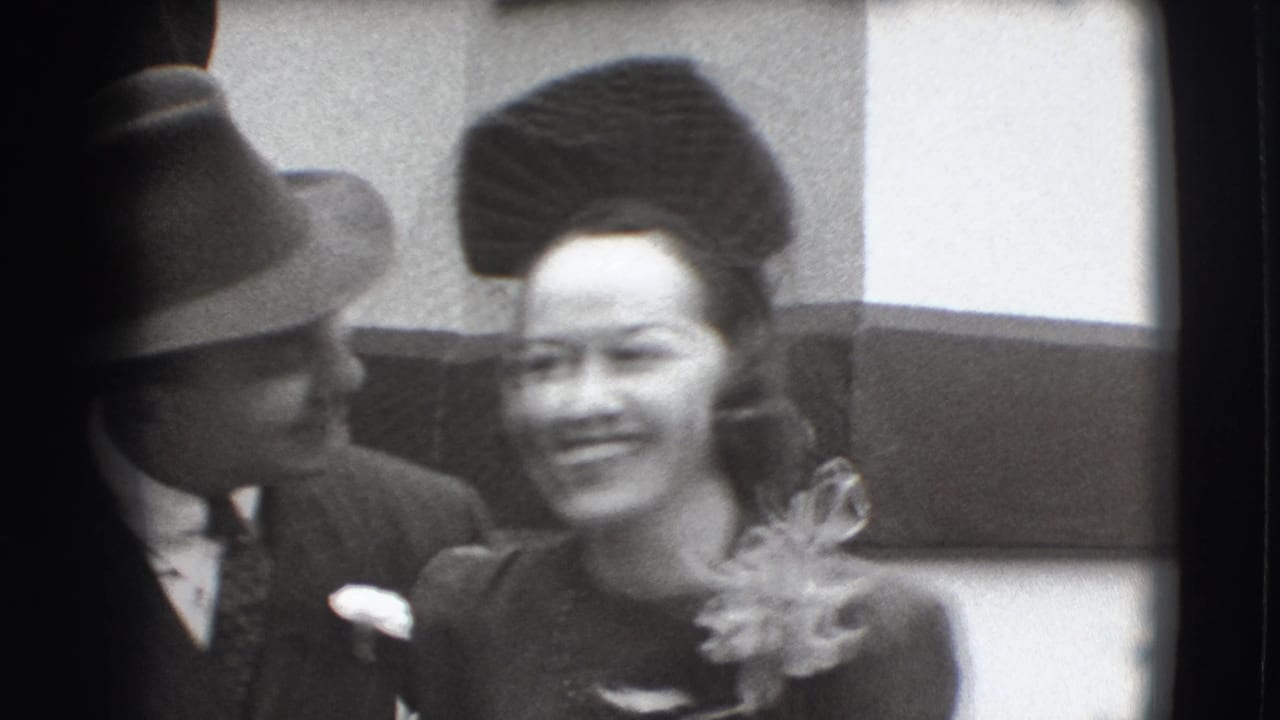Man in a fedora stands beside a smiling woman in a formal hat and dress.
