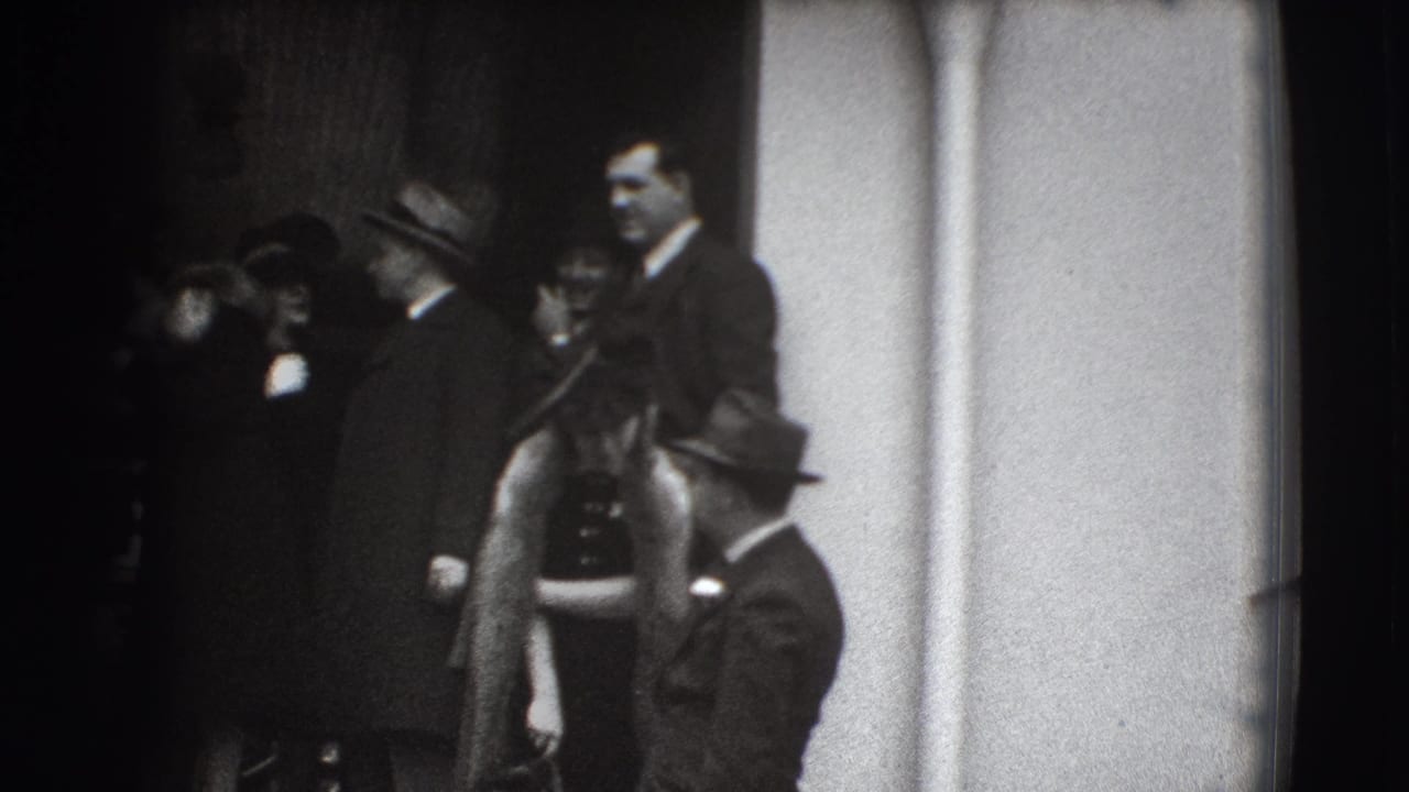 Formally dressed people stand near a building entrance in grainy black-and-white footage.