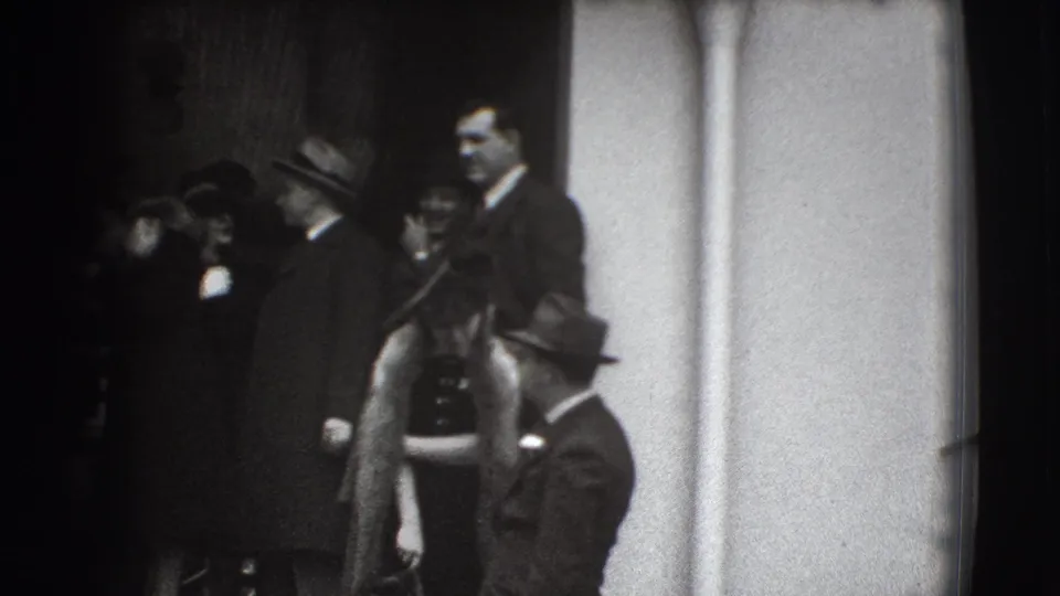 In Joliet, Illinois home-movie footage from 1941, suited men and women in hats gather near a doorway, partly obscured by soft focus.