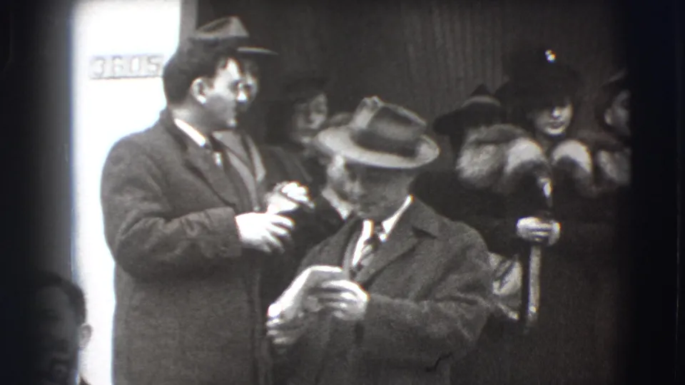 A suited man looks toward another man in a fedora as formally dressed women stand behind them in this 1941 Joliet home-movie frame.
