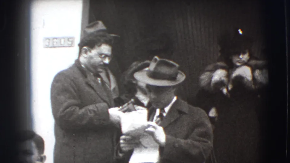 In 1941 Joliet, Illinois footage, two men in coats and hats stand close together with papers near a doorway and partial wall sign.