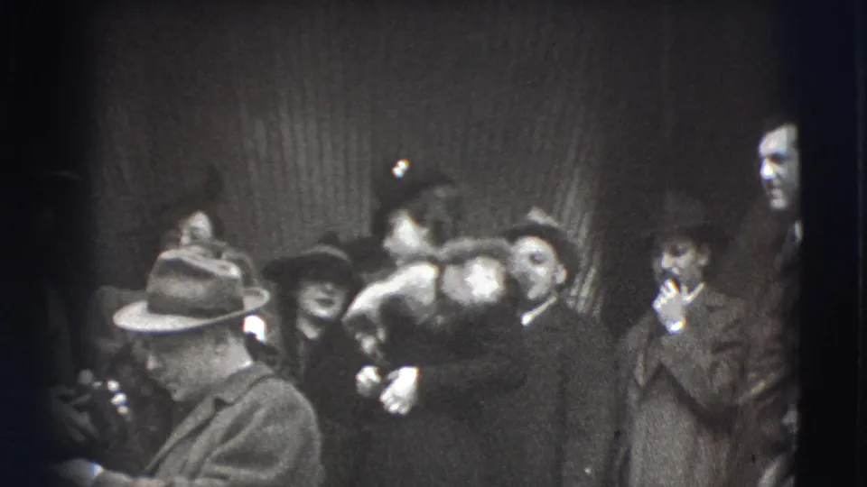 Adults in 1941 Joliet stand in a crowded line near a dark wall, including men in fedoras, women in coats, and a man smoking a pipe.
