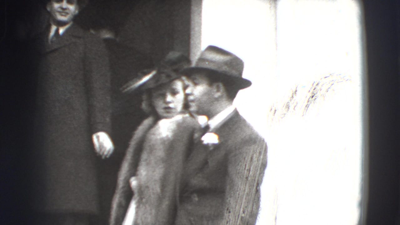 Adults in dress clothes stand near a bright window, including a woman in a fur-trimmed coat and men in hats.