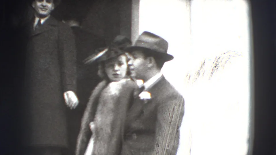 1941 Joliet home-movie frame showing adults in period dress near a bright interior window, with a fur-trimmed coat and fedoras visible.
