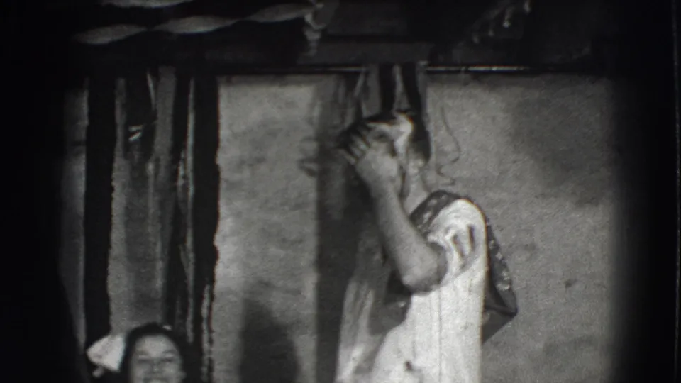 Black-and-white 8mm frame from the Joliet clip showing a man in a white shirt and loose tie raising his arms indoors.
