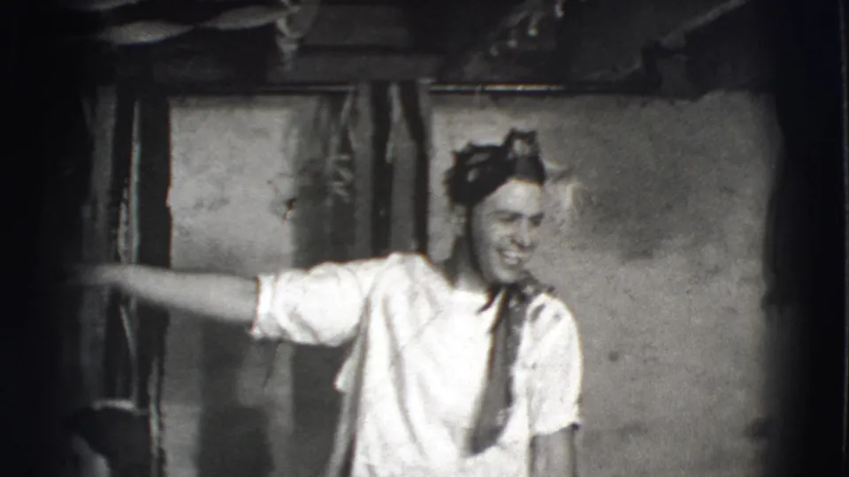 Black-and-white 8mm frame from the 1941 Joliet clip showing a smiling man in shirt and tie gesturing across an interior room.