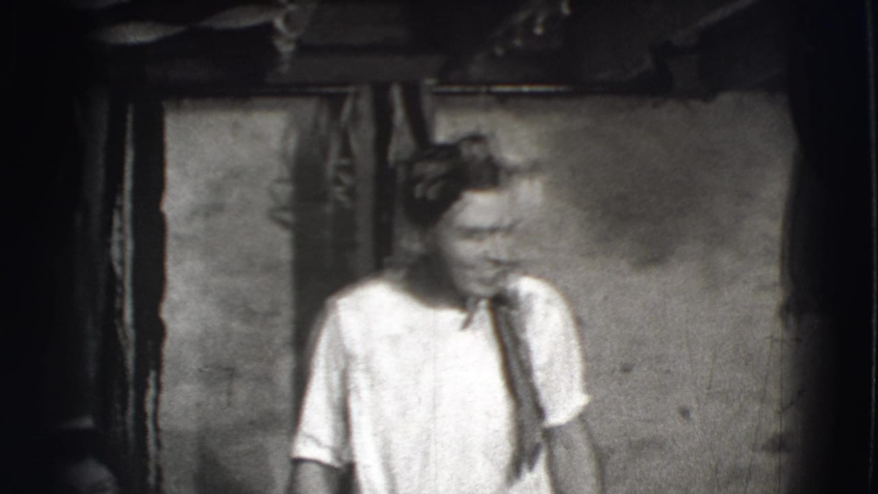 Man in a white shirt and tie stands in a dim interior with a headband or cloth on his head.